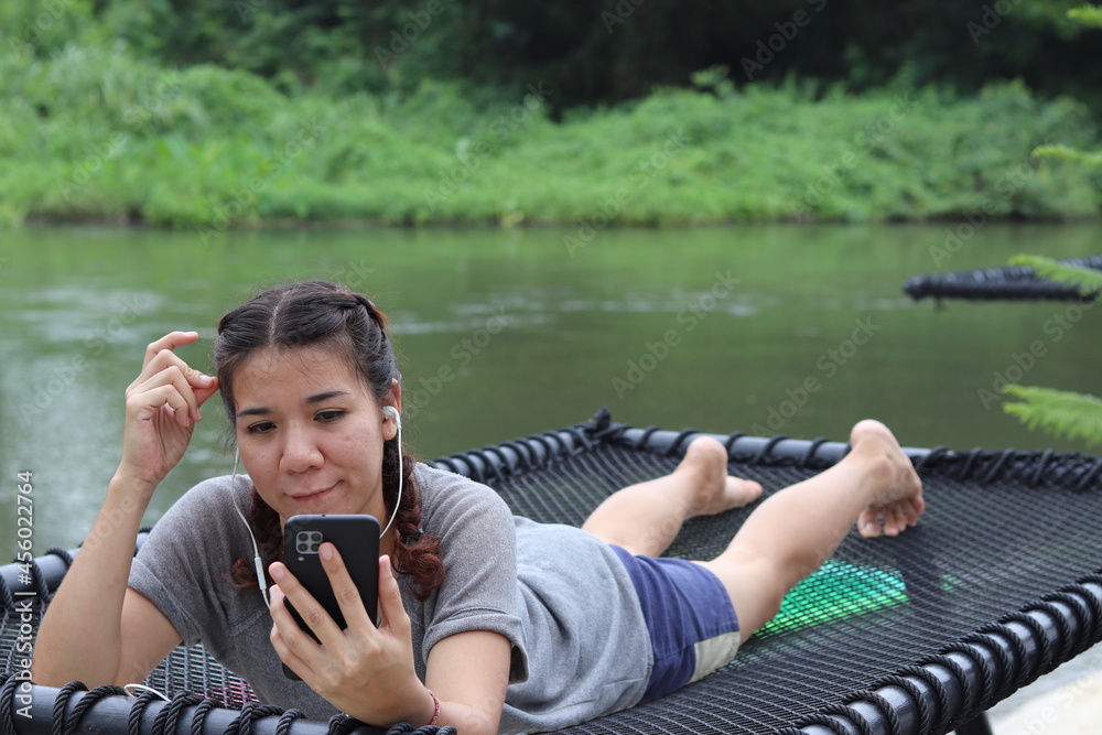 Fototapeta premium Asian woman tied hair white skin cute looking at mobile phone with headphones to play social media to relax on her vacation at the river Ideas for leisure activities, hobbies, stress relief, travel