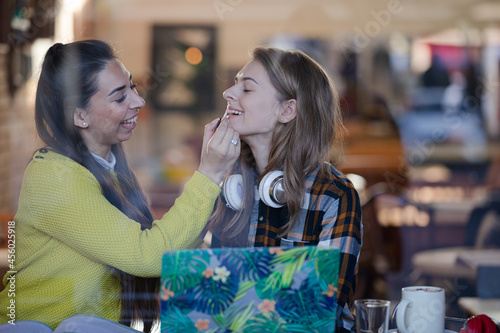 Young woman applying lip gloss to friends lips in cafe window