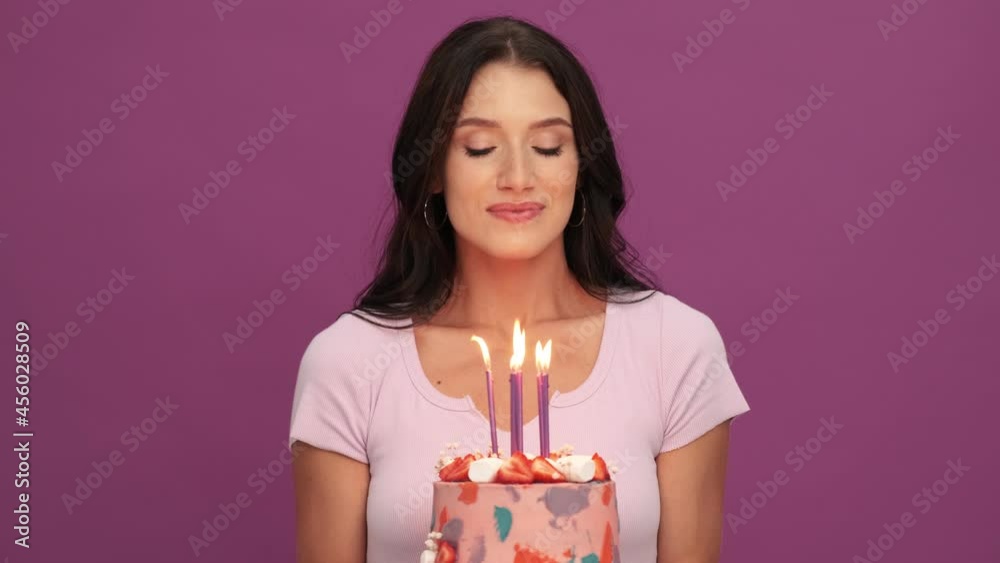 A smiling girl holding a birthday cake while making a wish and blow candles isolated over violet background in the studio A smiling girl holding a birthday cake while making a wish and blow candles isolated over violet background in the studio