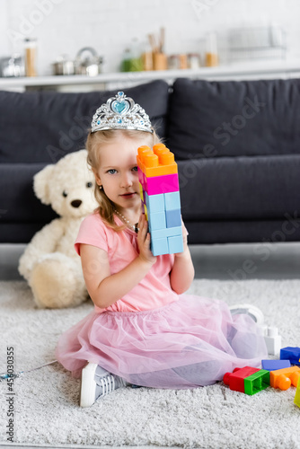 girl in toy crown showing tower of multicolored building blocks while sitting on floor