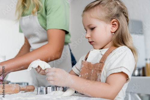 child holding dough near cookie cutters and blurred mom in kitchen