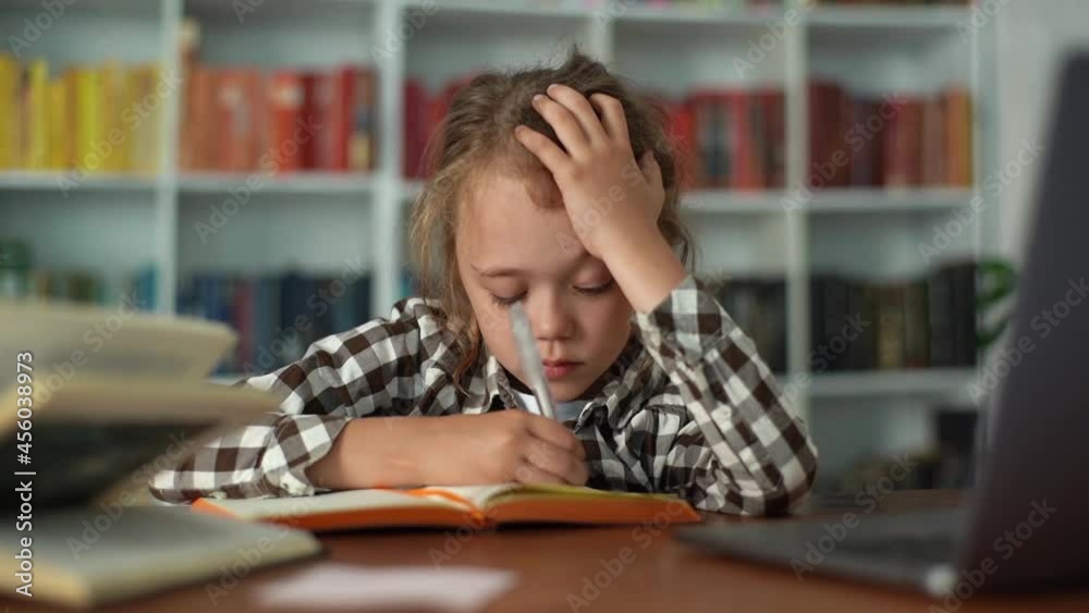 Front view of exhausted upset primary child school girl frustrated over homework holding head with hands. Tired pupil schoolgirl writing hometask in notebook and putting head on table, slow motion. Front view of exhausted upset primary child school girl frustrated over homework holding head with hands. Tired pupil schoolgirl writing hometask in notebook and putting head on table, slow motion.