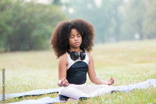 African American little girl doing meditate yoga asana on roll mat with eyes closed in park. Kid girl practicing doing yoga outdoor. Little afro girl with curly hairstyle training yoga