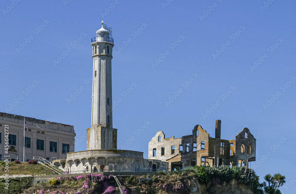USA, California, San Francisco - May 6, 2007: Alcatraz. Closeup of tall white lighthouse tower with ruins to the right and prison to the left under blue sky. Pink flowers add color.