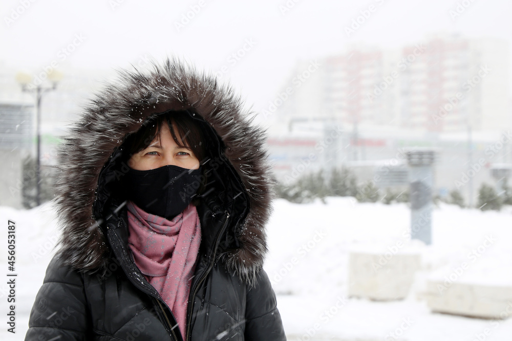Coronavirus protection at cold weather. Woman in black mask and fur hood standing on winter street during snow Coronavirus protection at cold weather. Woman in black mask and fur hood standing on winter street during snow