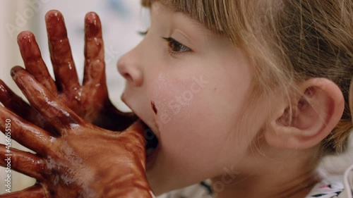 happy little girl with hands covered in chocolate licking fingers having fun baking in kitchen naughty child playing enjoying childhood