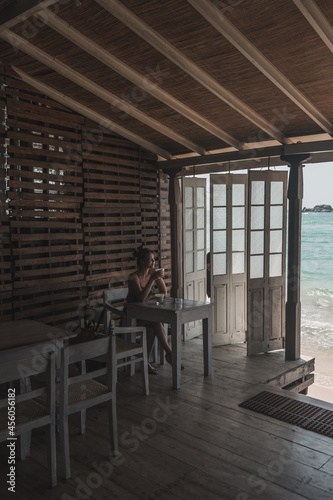 Girl sitting and drinking coffee in a wooden cafe overlooking the ocean. Blue crystal clear water in the ocean. Lovely view from the coffee shop.