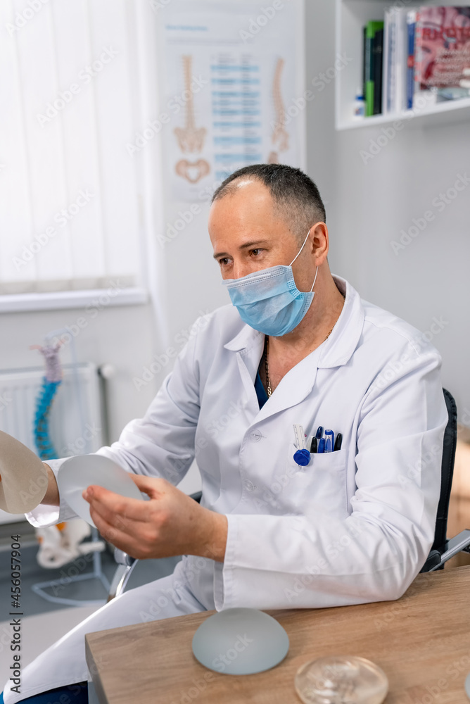 Doctor in mask showing silicon medical implants. Surgeon in medicine uniform holding new implants in hands. Stock photo
