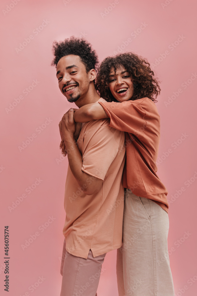 profile photo of young woman hugging from behind tall guy indoors in pink. happy brunettes with curly hair are dressed in light summer clothes. profile photo of young woman hugging from behind tall guy indoors in pink. happy brunettes with curly hair are dressed in light summer clothes.
