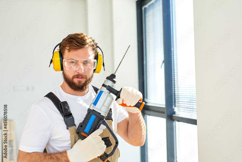 Fototapeta premium Young caucasian man holding power drill standing in front of white wall indoors,posing at camera, wearing overalls, headset and safety glasses. copy space, concept for home DIY