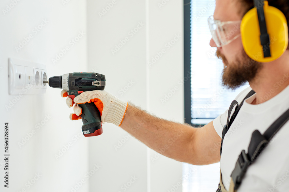 Side view portrait of young electrician setting the outlet in room on white wall, focused guy in headset and safety eyeglasses renovating room in new apartment. focus on hand with electric drill Side view portrait of young electrician setting the outlet in room on white wall, focused guy in headset and safety eyeglasses renovating room in new apartment. focus on hand with electric drill