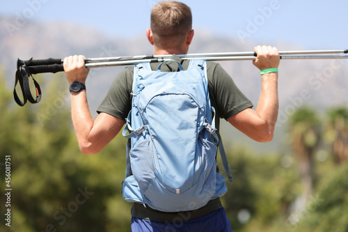 Man with backpack looking at mountains and holding walking sticks in his hands back view