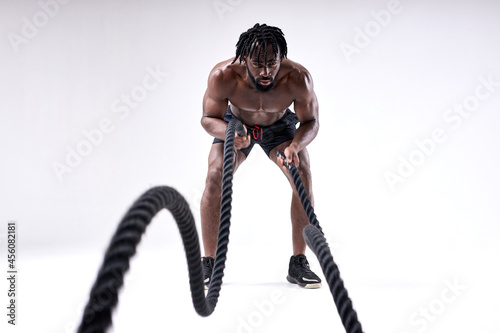 Sporty young black man doing cross fit exercises with ropes over isolated white background, training. hard workout. wave. shirtless fit athlete guy is concentrated on workout, sport. portrait