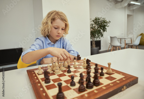 Organized caucasian boy looking pensive while playing chess, sitting at the table in school