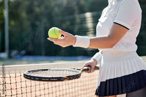 Close-up Photo Of Female Hands Tennis Player Holding Racket And Ball In Hands, Preparing For Game On Hardcourt, Standing By Net. Cropped Unrecognizabale Lady In Short Skirt And T-shirt Uniform © Roman