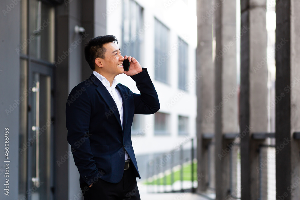 Asian businessman talking on the phone cheerful smiling reports good news near the office center outside Asian businessman talking on the phone cheerful smiling reports good news near the office center outside