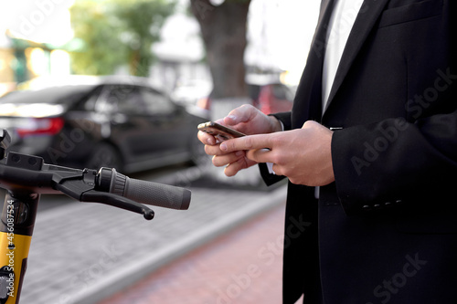 Cropped Man in formal suit paying for scooter rent through mobile application, stand with smartphone waiting. People lifestyle, modern transportation, vehicle. Close-up hands