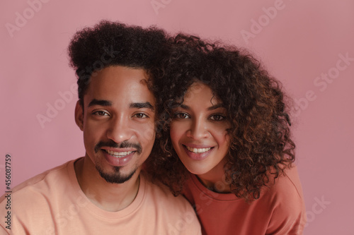 Cheerful dark-skinned guy with girl laughing while looking at camera on pink background. Curly brunettes are close together dressed in casual summer clothes. Good mood concept