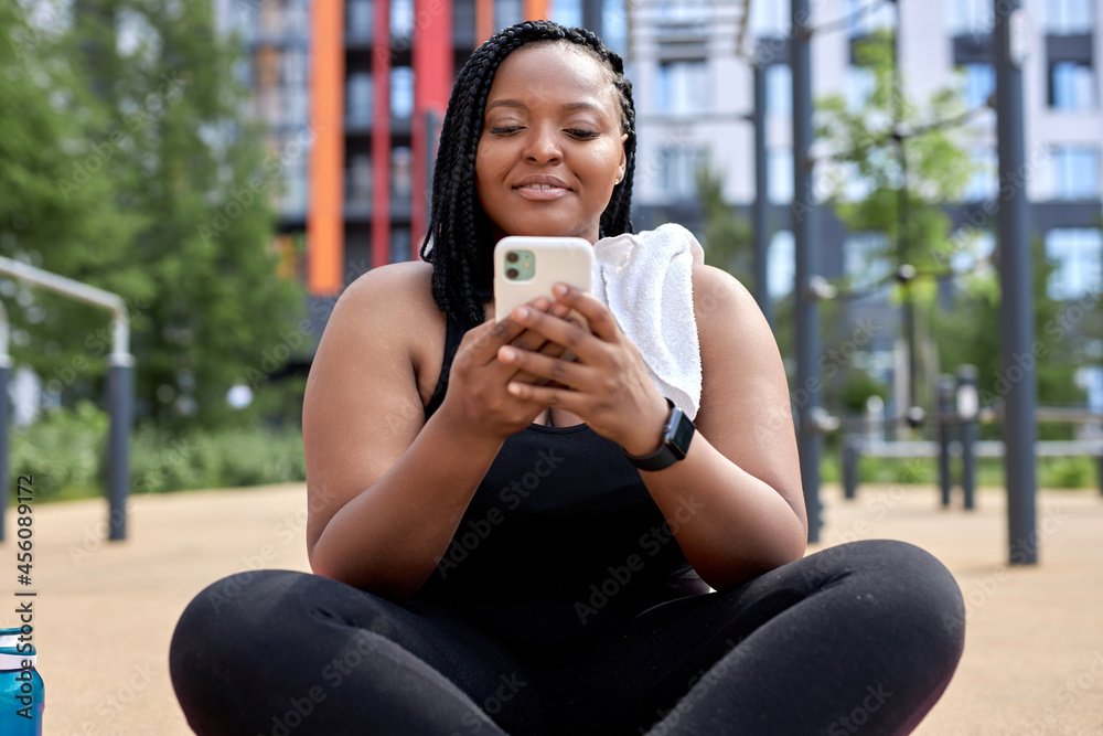 african woman sit in sports ground having rest chatting with friend on smartphone, typing message and smiling, enjoying sport, in black tracksuit, relax after workout. copy space. people lifestyle african woman sit in sports ground having rest chatting with friend on smartphone, typing message and smiling, enjoying sport, in black tracksuit, relax after workout. copy space. people lifestyle