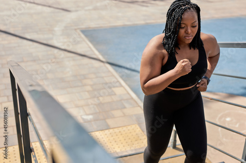 Obese young african woman foing fitness exercises on stairs outdoors. Weight loss cardio goal achievement challenge. Copy space. Side view portrait of black lady in black tracksuit engaged in fitness