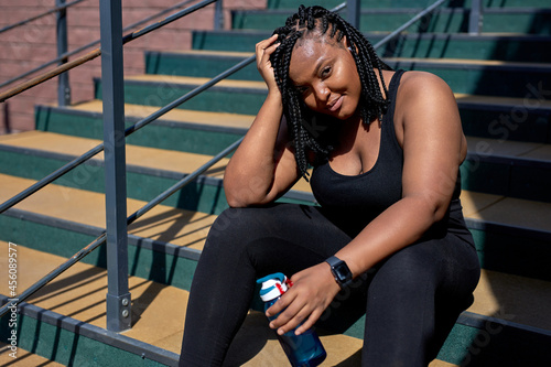 African Exhausted Female In Black Sportive Clothes Have Rest After Sport, Sitting With Water Bottle, Having Rest, Looking At Camera Smiling, Posing. In The Morning, Weight Loss, Sport, Fitness