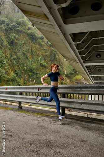 Side view on Young woman running on bridge in nature running for exercise. fitness, sport, people, exercising ,running and lifestyle. Side view portrait, copy space. Redhead lady in sportswear