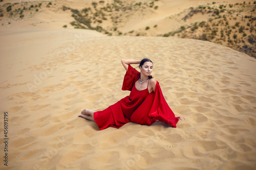 sexy brunette woman in a red long dress sit on a dune in the summer