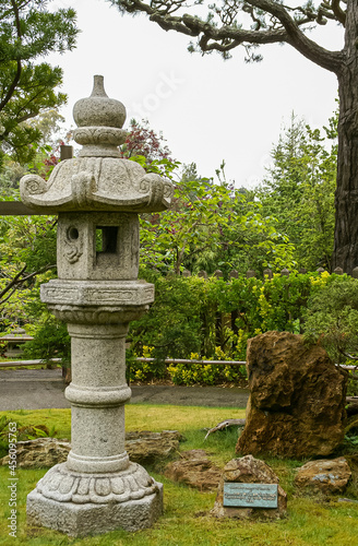 USA, California, San Francisco - May 19, 2008: Japanese Garden. Closeup of gray cut-stone lantern on lawn with different shades of green foliage and brown rock on side.