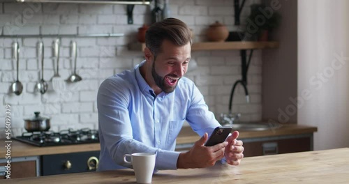 Focused businessman looking screen smartphone says yes and makes a hand gesture expressing victory. Male sit at the table kitchen background. Winning concept