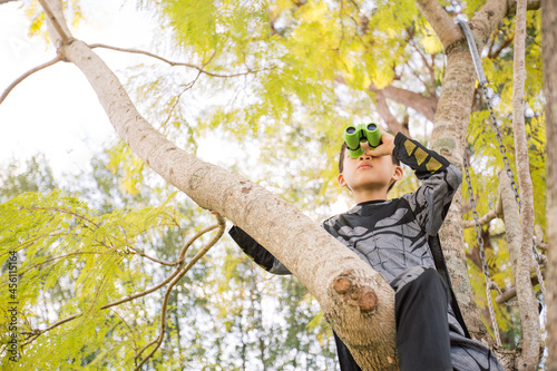 Little batman boy climbing a tree in the garden looking through a binocular