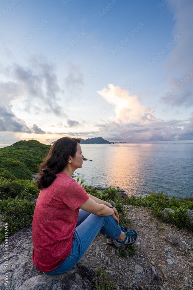 A woman sits on mountains and looks at the ocean. 