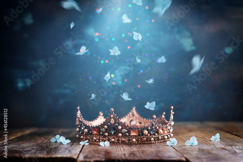 low key image of beautiful queen or king crown over wooden old table and falling flowers. fantasy medieval period