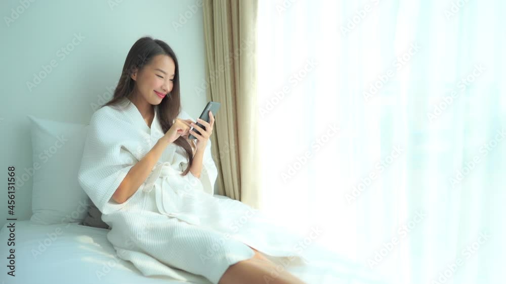 Woman relaxing on bed using phone, smiling as she scrolls seated near window with morning light. Asian lady in white robe lounging in luxury resort bedroom looking at smartphone. Woman relaxing on bed using phone, smiling as she scrolls seated near window with morning light. Asian lady in white robe lounging in luxury resort bedroom looking at smartphone.