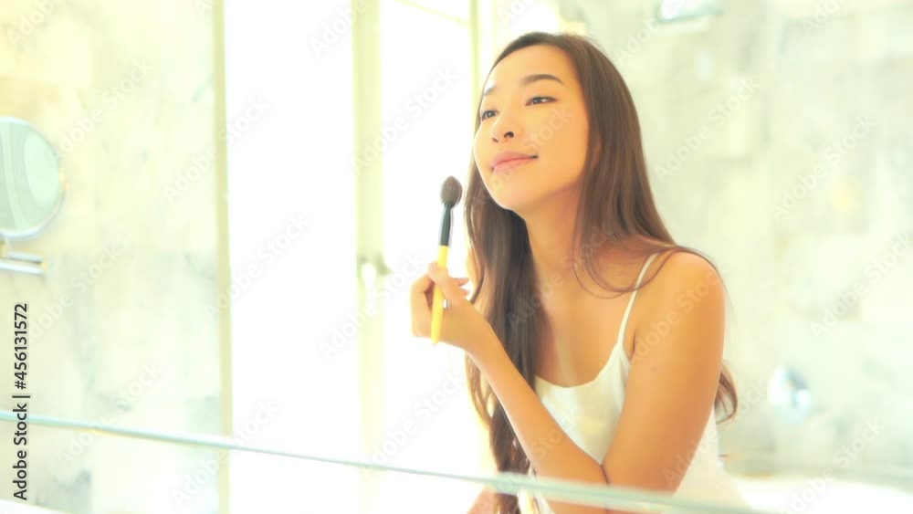 Asian female applies skin powder on the face with a brush in her bathroom, reflection in the mirror Asian female applies skin powder on the face with a brush in her bathroom, reflection in the mirror