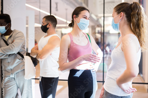 Young girls in sportswear and protective face masks talking friendly in modern fitness studio hall after group pilates workout. New life reality in pandemic