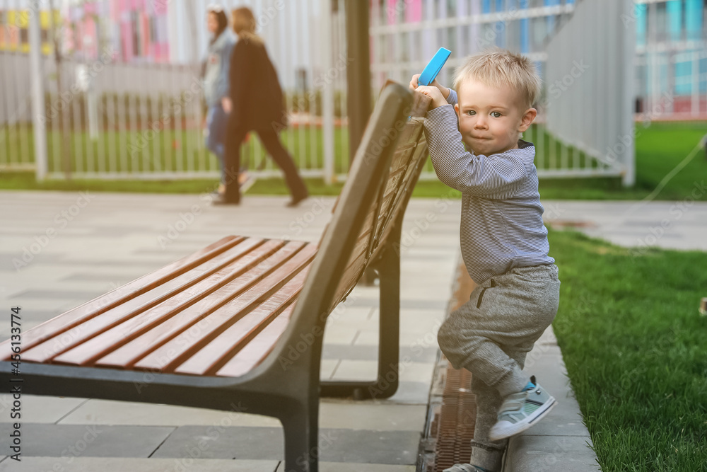 Pensive boy with blond hair with a shovel sits on a bench in park on the background of apartment houses and women Pensive boy with blond hair with a shovel sits on a bench in park on the background of apartment houses and women