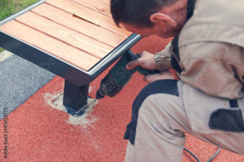 Blur image of installation of benches on the playground. Bench and holes on artificial grass for fitting, mounting. A man in overalls drills a hole with a drill for a screw.