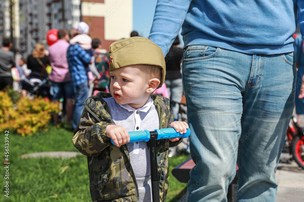 Daddy rolls his son on a scooter. A child in a military cap on the parade of the day of victory in Russia against the backdrop of a crowd