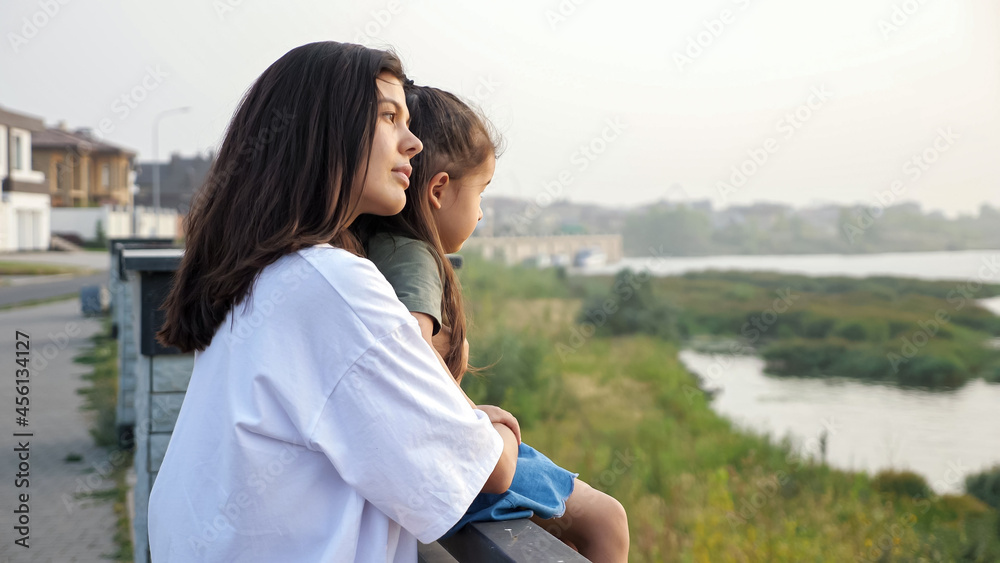 Careful mother holds hand of Asian little girl sitting on large parapet pedestal on contemporary city embankment at sunset, side view