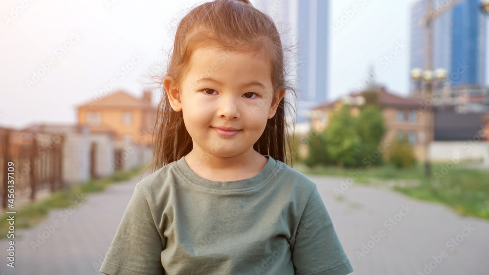 Adorable happy little Asian girl in green t-shirt poses for camera standing on city waterfront against buildings in summer evening close view, sunlight