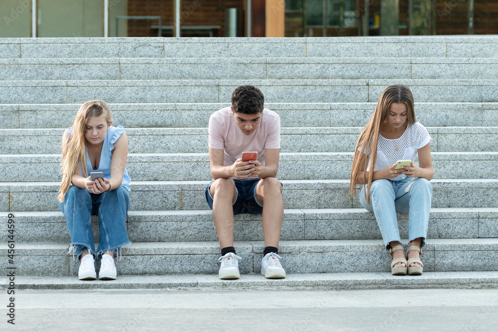Digital addiction on generation Z teenagers concept. Three friends sitting on stairs concentrated and chatting on their phones.