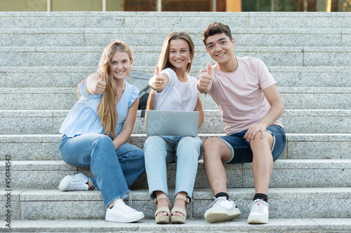 Successful college students gesturing thumbs up or making the ok sign. Happy teenagers sitting on stairs. Teamwork in adolescence concept.