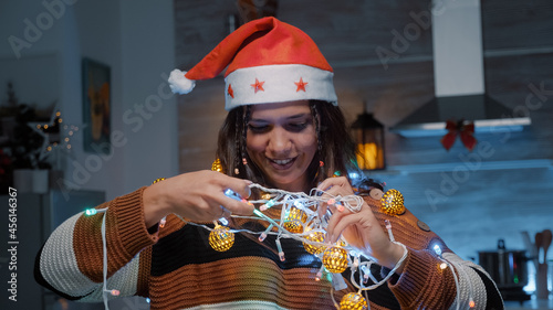 Festive woman with santa hat trying to untangle lights while decorating kitchen for christmas eve festivity. Young adult holding knotted garland for tree and preparing for dinner celebration photo