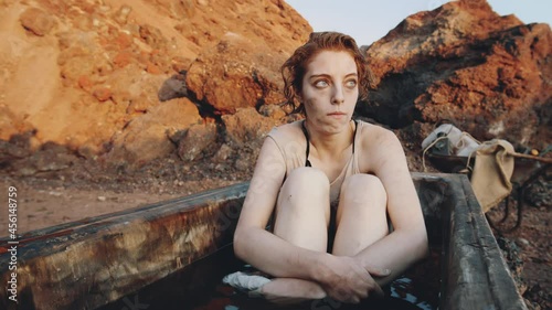 Young shivering woman in wet worn clothes sitting in old dirty bath and looking in distance in post apocalyptic world