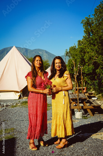 Full length portrait of mother and daughter holding flowers while standing against tents at campsite on sunny day