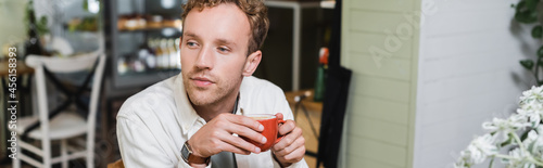 curly man holding cup of cappuccino and looking away in cafe, banner