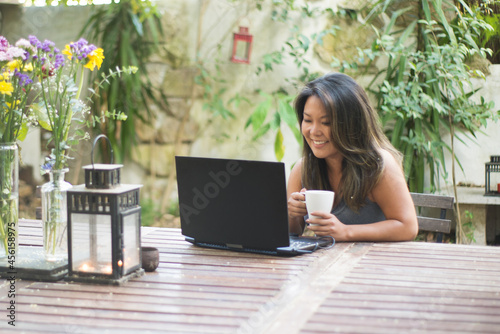 Portrait of attractive Japanese woman using laptop at backyard. Beautiful girl shopping or chatting online, having fun, watching movie, freelancer working. Drinking coffee. Modern technology concept