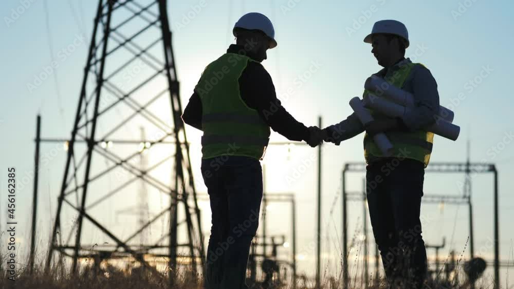 Handshake of two men people work industry. Engineers shaking hands at the construction of high-voltage power. Team engineers looking plan. Silhouette two engineer standing on field at sunset time.