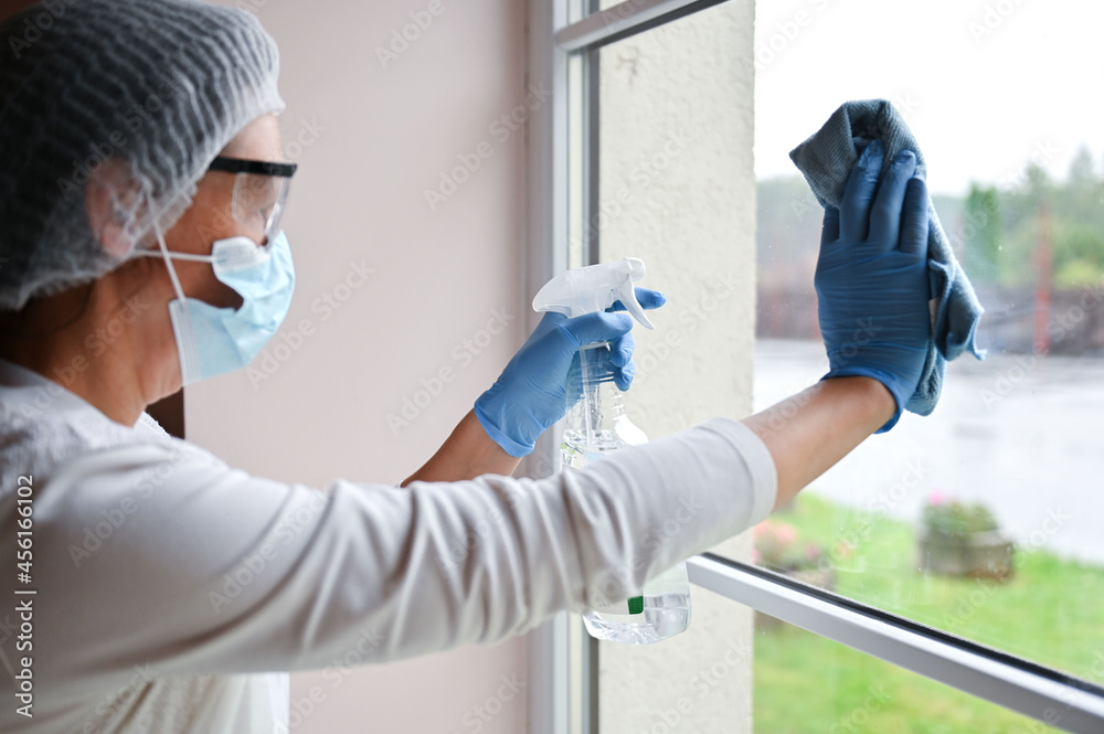 Naklejka premium Mature woman in protective medical suit and respirator mask desinfecting furniture and cleaning window glass at hospital