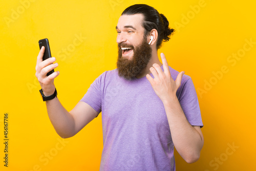 Excited bearded man is using his earphones and phone over yellow background.